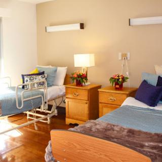 A well-lit hospital room featuring two beds and bedside furniture.