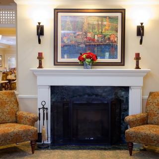 A cozy living room with a fireplace flanked by two armchairs, adorned with a painting and flowers.