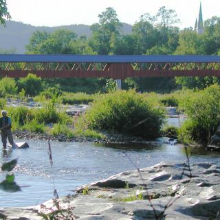A peaceful river scene with a person fishing near a picturesque covered bridge.