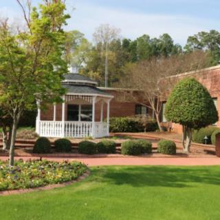 A beautiful garden with a white gazebo, neatly trimmed shrubs, and a brick walkway adjacent to a red brick building.