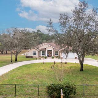A suburban house with a circular driveway, surrounded by trees and a fenced yard under a partly cloudy sky.