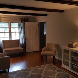 A cozy living room with hardwood floors, featuring armchairs, a rug, and large windows with curtains.