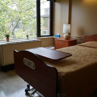A patient room featuring a bed, bedside table, lamp, and window with a view of greenery.