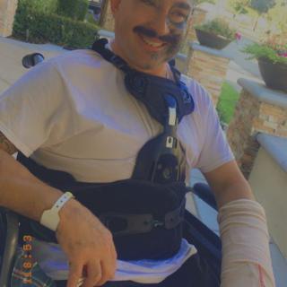 A man in a wheelchair with a cast on his arm, smiling outdoors.