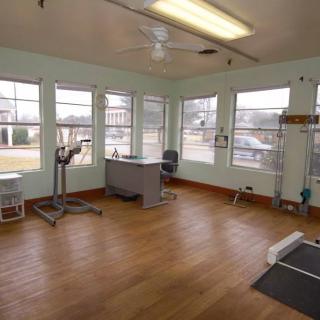 An empty physical therapy room with equipment, a desk, and large windows providing natural light.
