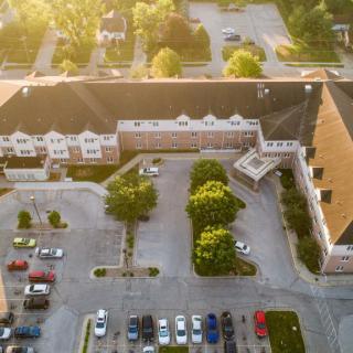 An aerial view of a building with a parking lot and surrounding trees under soft sunlight.