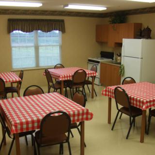 A dining area with multiple tables covered in red checkered tablecloths, surrounded by brown chairs. The room includes a small kitchen space with a refrigerator and microwave.