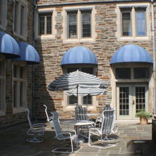 A quaint courtyard featuring outdoor seating with a striped umbrella, surrounded by a stone building with blue awnings.