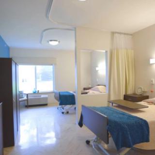 A bright and clean hospital room with two patient beds separated by a curtain, featuring large windows.