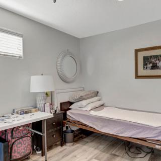A minimalist bedroom with a single bed, featuring gray walls and wooden flooring, is simply decorated with a lamp, mirror, and framed picture.