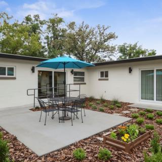 A cozy outdoor patio area featuring a table with chairs under a blue umbrella, surrounded by lush garden landscaping.