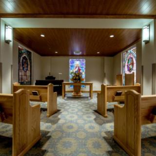 A serene chapel interior featuring wooden pews and a stained glass window at the altar.