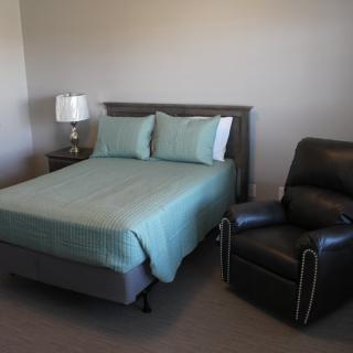 A cozy bedroom with a neatly made bed, a recliner chair, and a nightstand with a lamp.