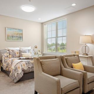 A cozy and well-decorated bedroom with neutral tones, featuring a bed, two armchairs, and a table lamp by the window.