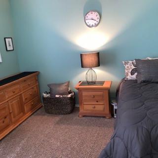 A cozy bedroom featuring a neatly arranged nightstand with a lamp, dresser, and a clock on the teal wall.