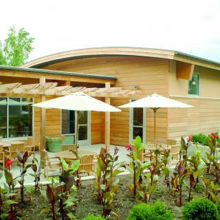 A modern wooden building with a patio featuring umbrellas and landscaped surroundings.