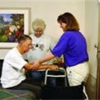A caregiver assists an elderly man with physical therapy while another person observes.