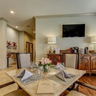 A well-decorated dining room with a table set for four, complete with napkins and floral centerpiece, complemented by elegant furnishings and a wall-mounted television.