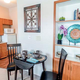 A cozy dining area adjacent to a small kitchen, featuring a table set for two and decorative shelving with plants and artwork.