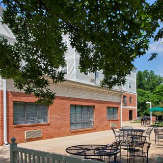 An outdoor patio with tables and chairs next to a brick building under a tree on a sunny day.