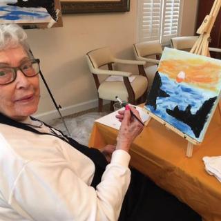 An elderly woman painting a scenic landscape on a canvas indoors.