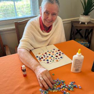An elderly woman enjoys creating a colorful mosaic art piece using various craft materials.