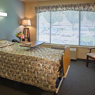 A well-lit hospital room featuring a bed, chair, lamp, and large window with floral curtains.