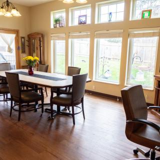 A well-lit dining room with a large table and chairs, surrounded by tall windows.