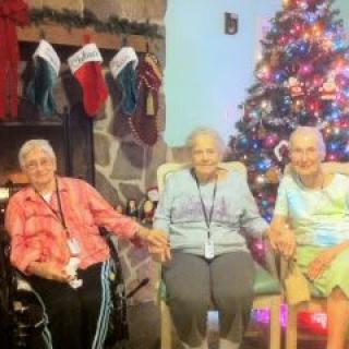 Three elderly people sitting in front of a decorated Christmas tree and fireplace, enjoying the holiday season together.