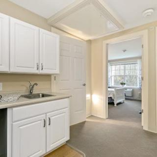 A small kitchenette with white cabinets and a sink, leading to a bedroom with ample natural light.