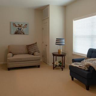 A cozy living room with a sofa, chair, and a side table featuring a lamp, complemented by wall artwork and natural light from the window.