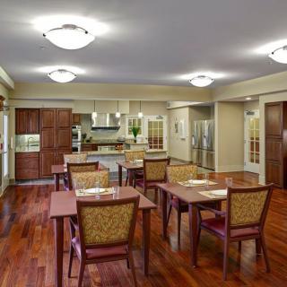 A spacious and modern dining area with wooden flooring and well-arranged tables and chairs, adjacent to a kitchen.