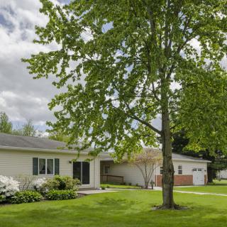 A suburban house with a grassy yard and a large tree in the foreground under a cloudy sky.