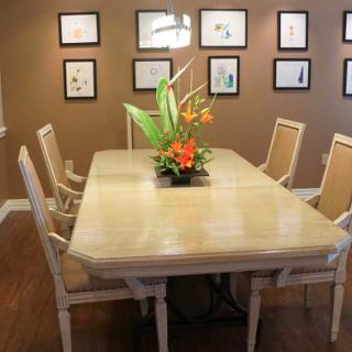 A modern dining room with a large table, surrounded by chairs and adorned with a central floral arrangement. The walls are decorated with framed artwork.
