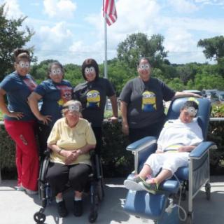 A group of caregivers and elderly individuals sit and stand together outdoors near an American flag.