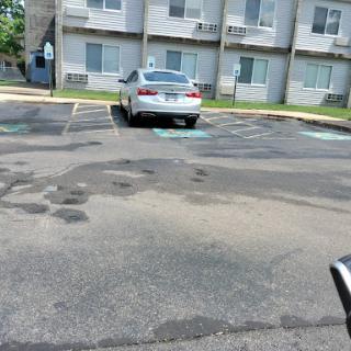 A white car parked in an apartment complex parking lot with visible asphalt patches.