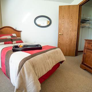 A cozy bedroom with a neatly made bed featuring a striped comforter, an oval mirror on the wall, and wooden furniture including a dresser and open door closet.