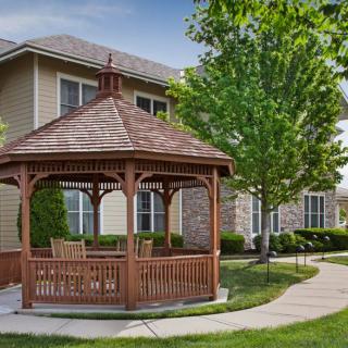 A wooden gazebo surrounded by a landscaped lawn and trees, with a house in the background.