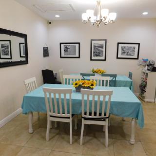A well-lit dining room with tables covered in blue tablecloths, adorned with flower arrangements and surrounded by white chairs, showcasing art on the walls and a large mirror.