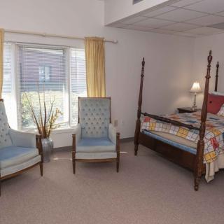A cozy bedroom featuring a four-poster bed, two upholstered chairs, and a large window with yellow curtains.