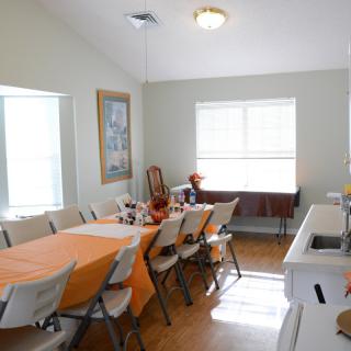 A dining room with a long table covered in an orange tablecloth, surrounded by folding chairs and lit by natural light from the windows.