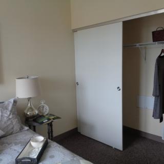 A bedroom with an open closet showing hanging clothes and a bedside table with a lamp.