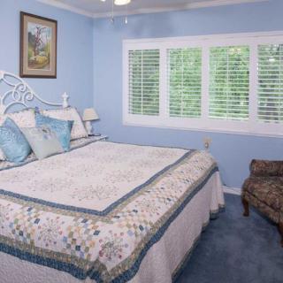 A cozy bedroom with soft blue walls, featuring a neatly made bed with a quilt and pillows. The room has a window with white blinds and a patterned chair in the corner.