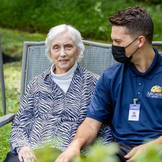 An elderly woman and a caregiver sit together outdoors, engaged in conversation.