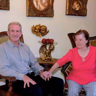 An elderly couple sitting indoors, holding hands and surrounded by artwork.
