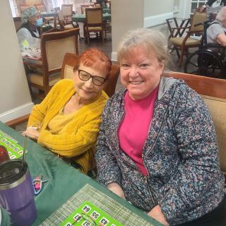 Two women are seated at a table, smiling in a social setting.