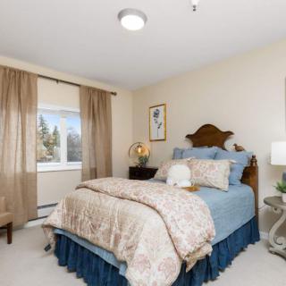 A cozy bedroom with a neatly made bed, decorated with elegant bedding and flanked by bedside tables holding lamps. The room features large windows with flowing curtains, allowing natural light to filter in.