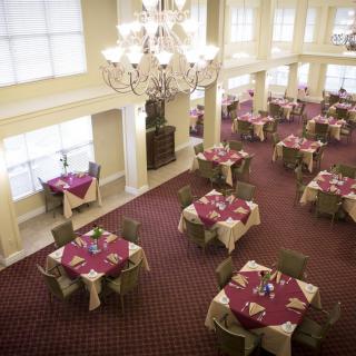An elegant dining area featuring multiple tables with red tablecloths, surrounded by chairs, under a large chandelier.
