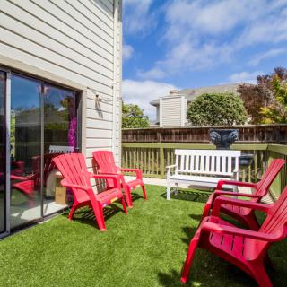 A sunny backyard patio with red chairs and a white bench on green artificial grass.