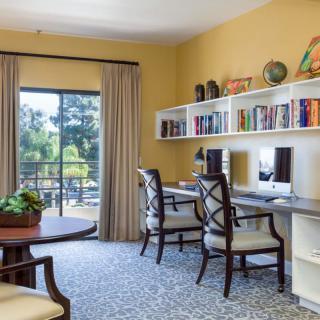 A cozy home office with dual computer workstations, bookshelves, and a view outside.